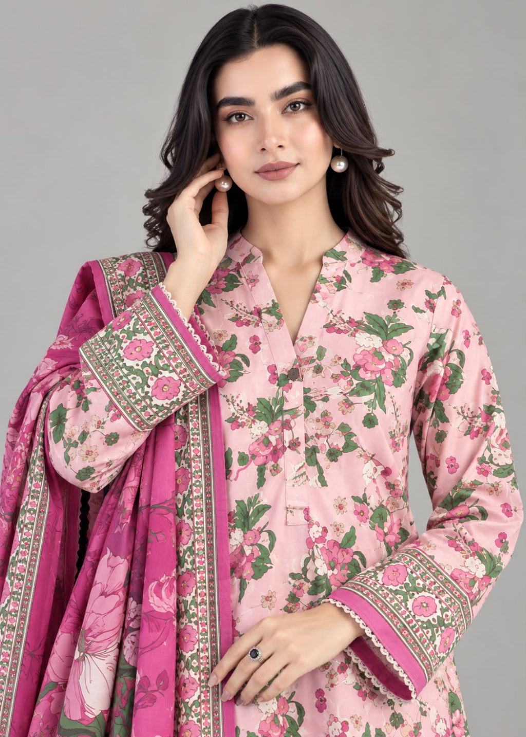 A woman modeling a pink floral traditional outfit with a matching pink dupatta, featuring green leaves and detailed gold and white border embroidery, set against a solid gray studio background.