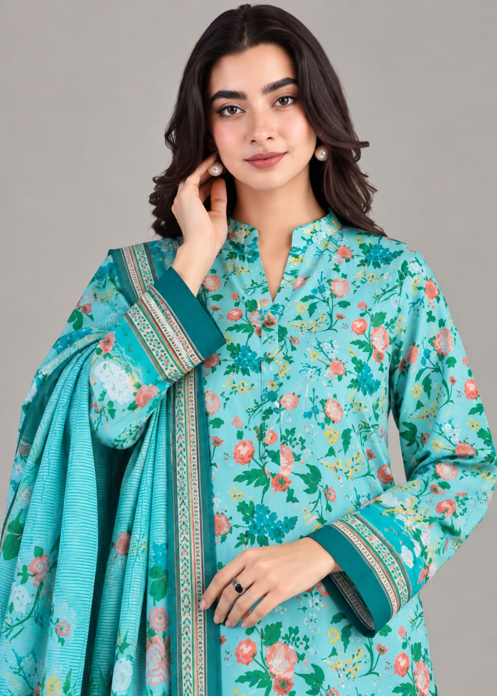 A woman modeling a light turquoise floral salwar kameez suit with a matching dupatta, featuring green and pink floral prints and gold patterned borders on the cuffs and collar, set against a solid gray studio background.