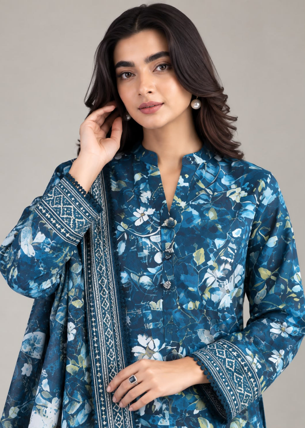 A woman modeling a blue floral printed kurta with a matching dupatta, featuring intricate geometric border patterns on the sleeves and collar, set against a solid gray studio background.