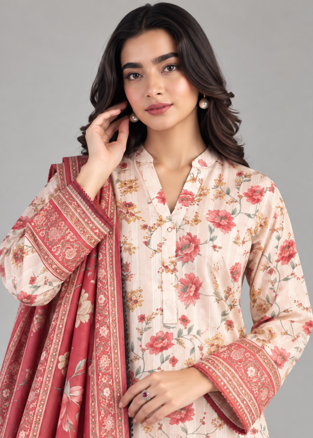 A woman modeling a traditional floral printed kurta in a light beige-pink with red and green floral motifs, paired with a matching red dupatta featuring ornate gold borders, set against a solid gray studio background.