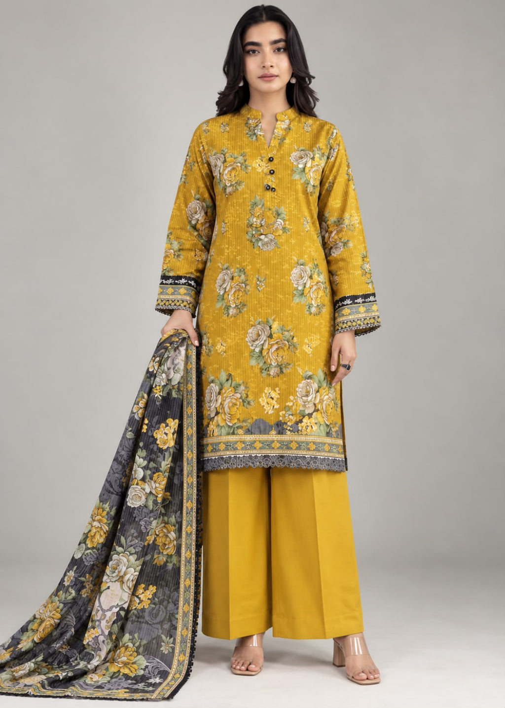A woman modeling a traditional Indian outfit consisting of a mustard yellow floral printed kurta, matching wide-leg trousers, and a dark gray dupatta with yellow and white floral motifs, set against a plain light gray studio background.