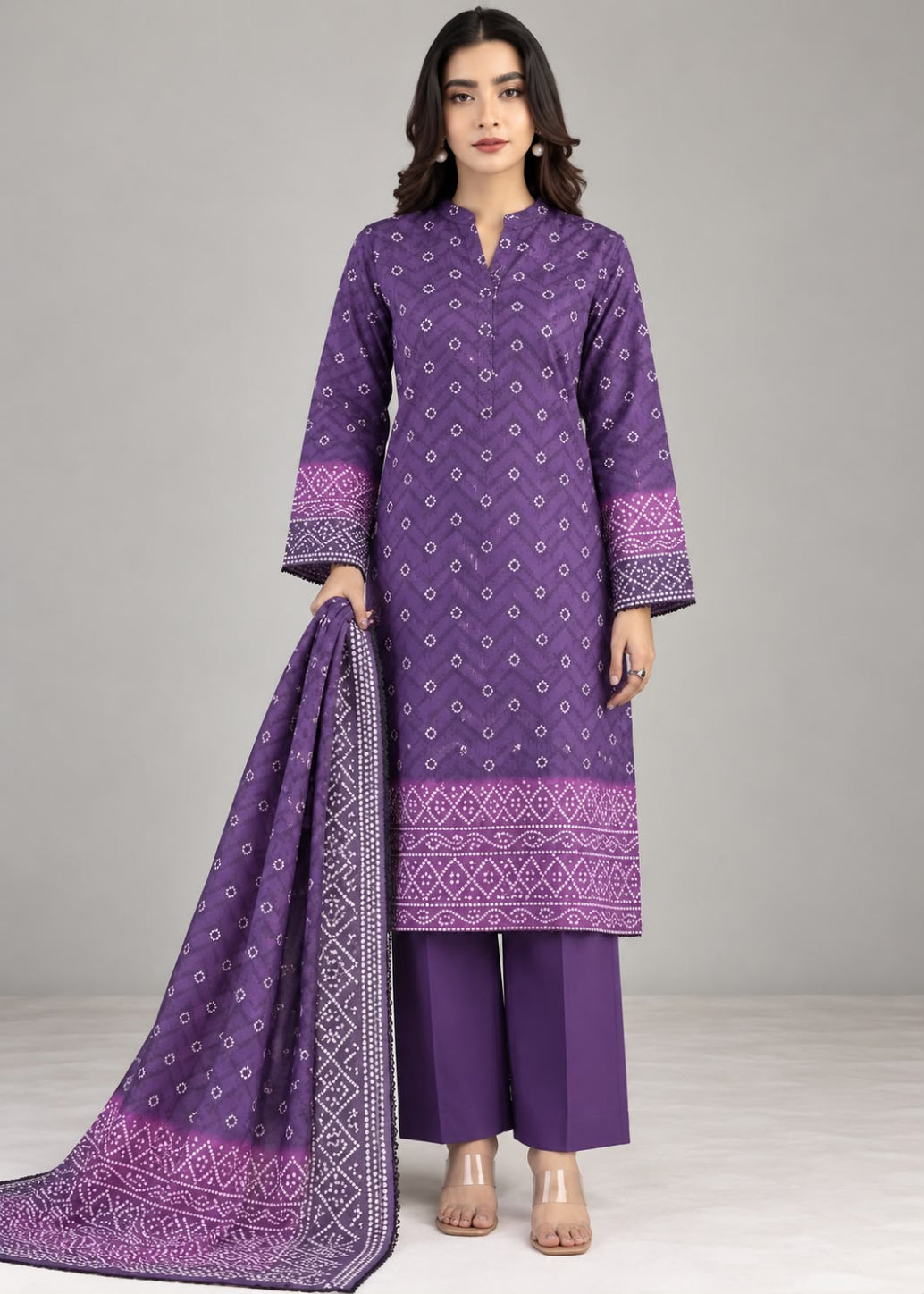 A woman modeling a purple three-piece traditional outfit with matching printed dupatta, featuring a kurta with white geometric patterns, wide-leg trousers, and a coordinating patterned scarf against a neutral gray studio background.
