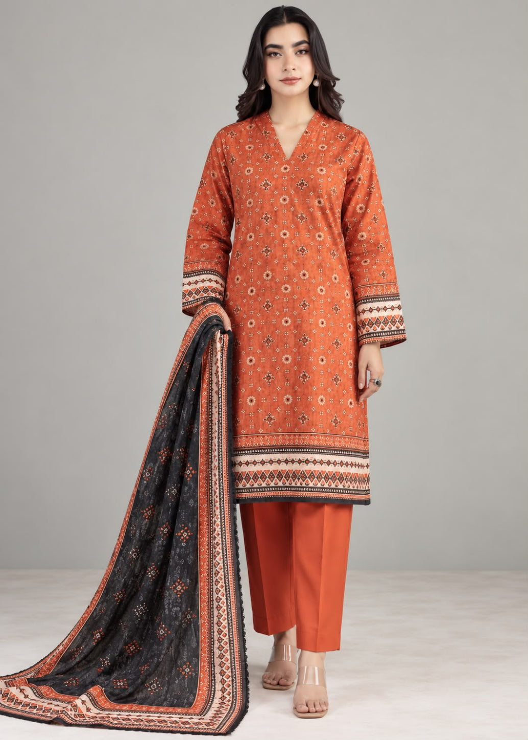 A woman modeling a traditional three-piece salwar kameez suit. The set features an orange kurta with intricate white and black geometric prints, matching orange trousers, and a black dupatta with a detailed orange and white border.