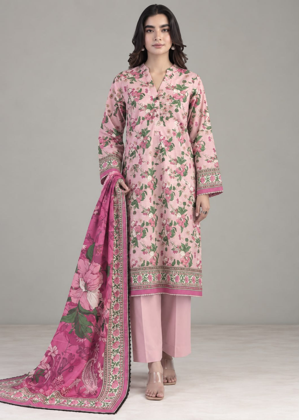 A woman models a three-piece pink ethnic suit set featuring a floral printed kurta, solid pink pants, and a matching pink floral dupatta with a decorative border, set against a neutral gray studio background.