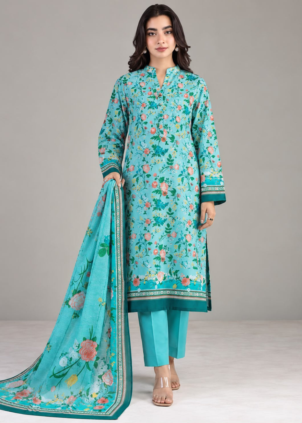 A woman modeling a traditional three-piece salwar kameez suit in turquoise blue, featuring a floral print, matching trousers, and a coordinating dupatta, set against a neutral studio background.