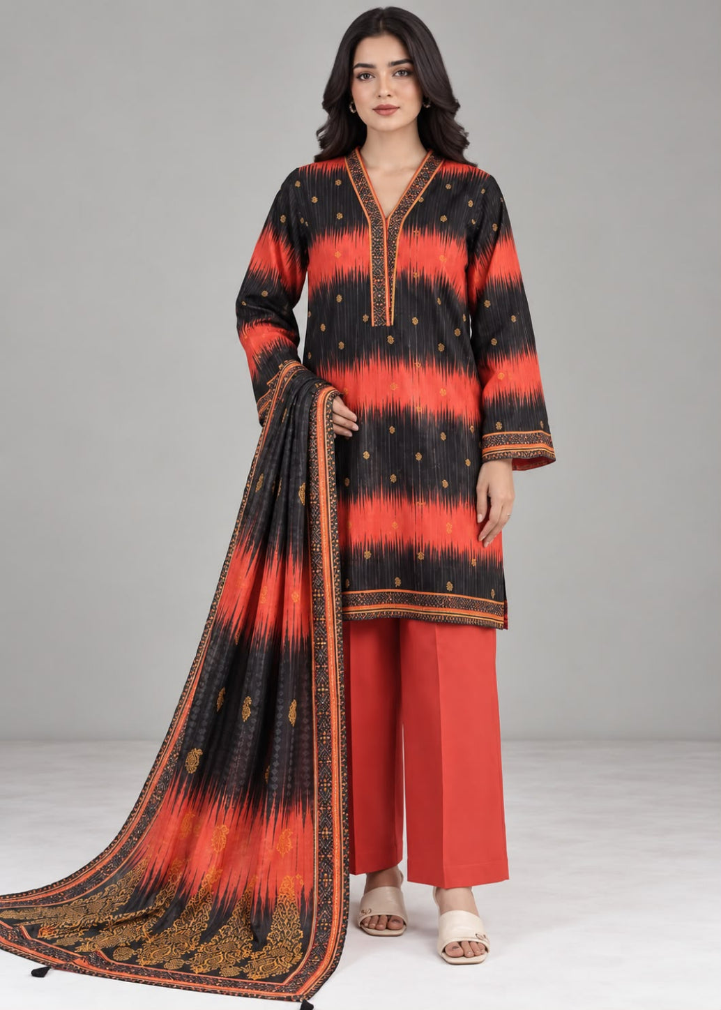 A woman modeling a traditional Indian outfit consisting of a black and orange tie-dye kurta with gold accents, matching orange trousers, and a coordinating dupatta, set against a plain gray studio background.