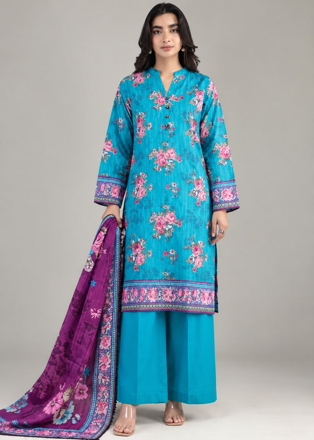 A woman models a traditional three-piece salwar kameez suit set. The set features a turquoise blue printed kurta with pink floral motifs and a purple border, matching wide-leg trousers in turquoise, and a purple dupatta with pink floral prints and a blue border.