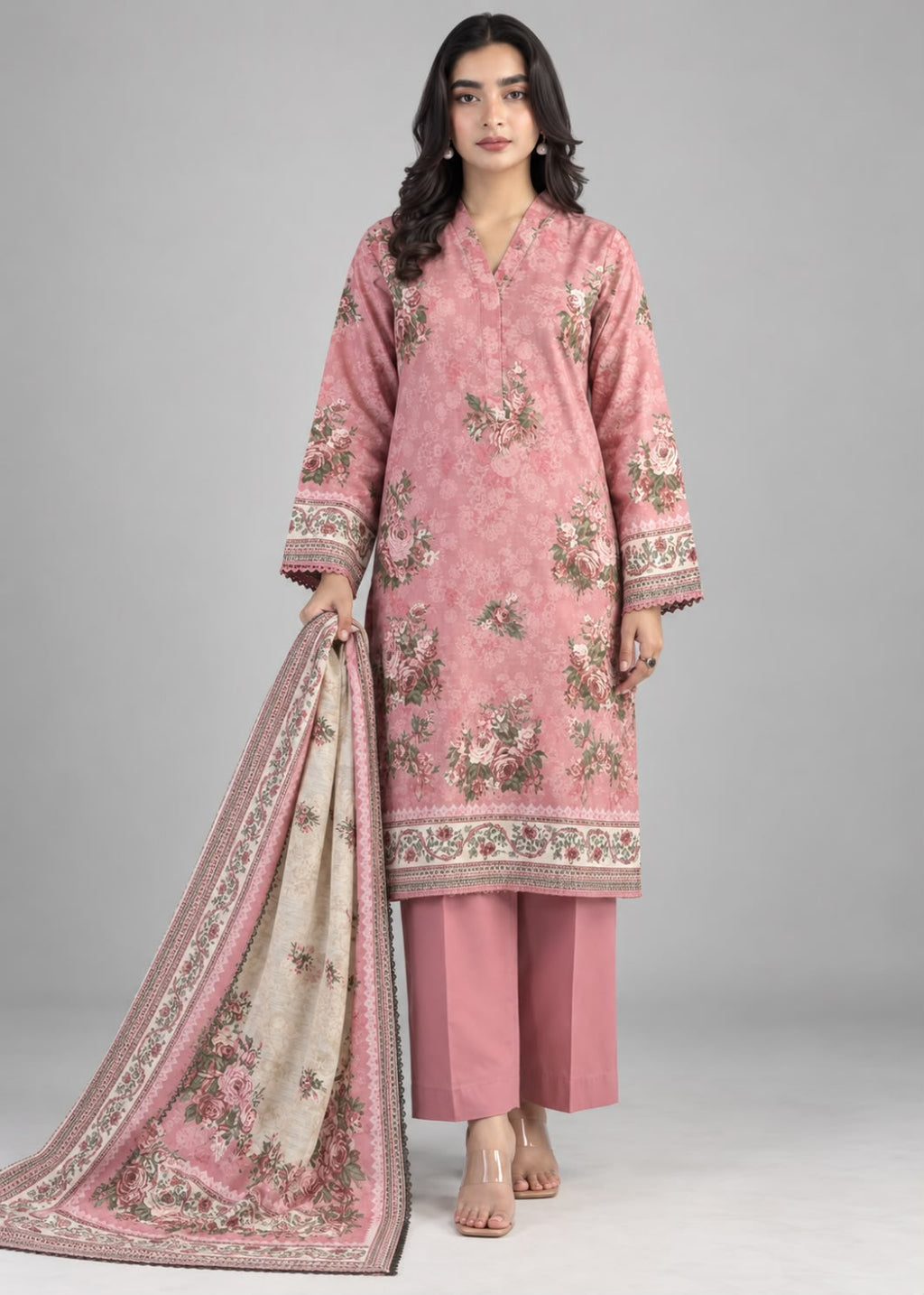 A woman modeling a pink traditional three-piece suit set with floral embroidery, matching trousers, and a beige dupatta with pink floral borders, photographed against a neutral gray studio background.