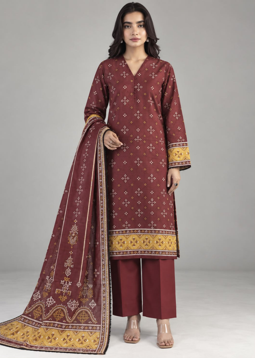 A woman modeling a traditional three-piece salwar kameez suit in maroon with gold and white printed patterns, paired with matching maroon trousers and a coordinating dupatta, set against a plain gray studio background.