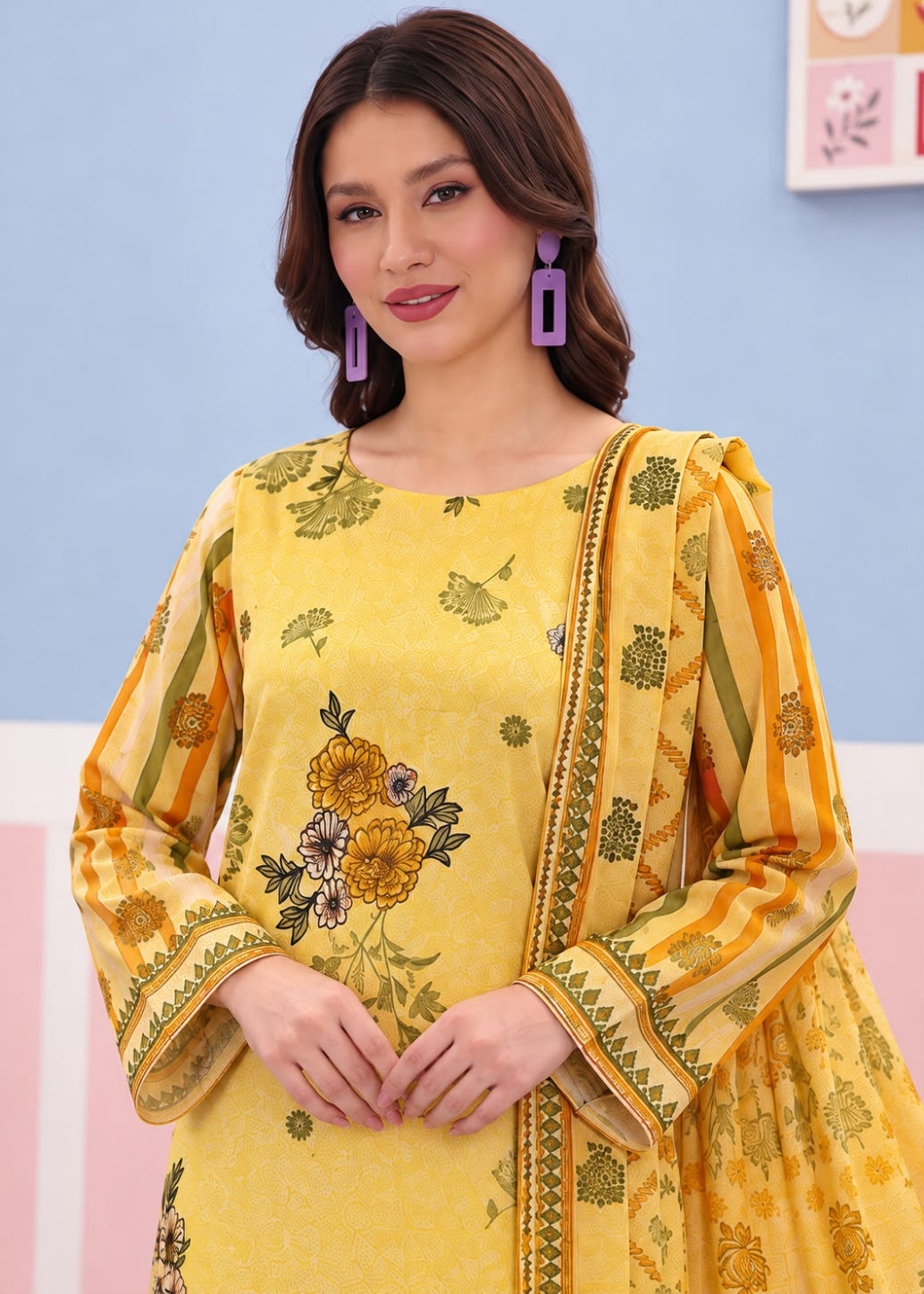 A woman wearing a yellow printed traditional outfit with a matching dupatta, featuring floral embroidery and green motifs, set against a pastel blue and pink background.