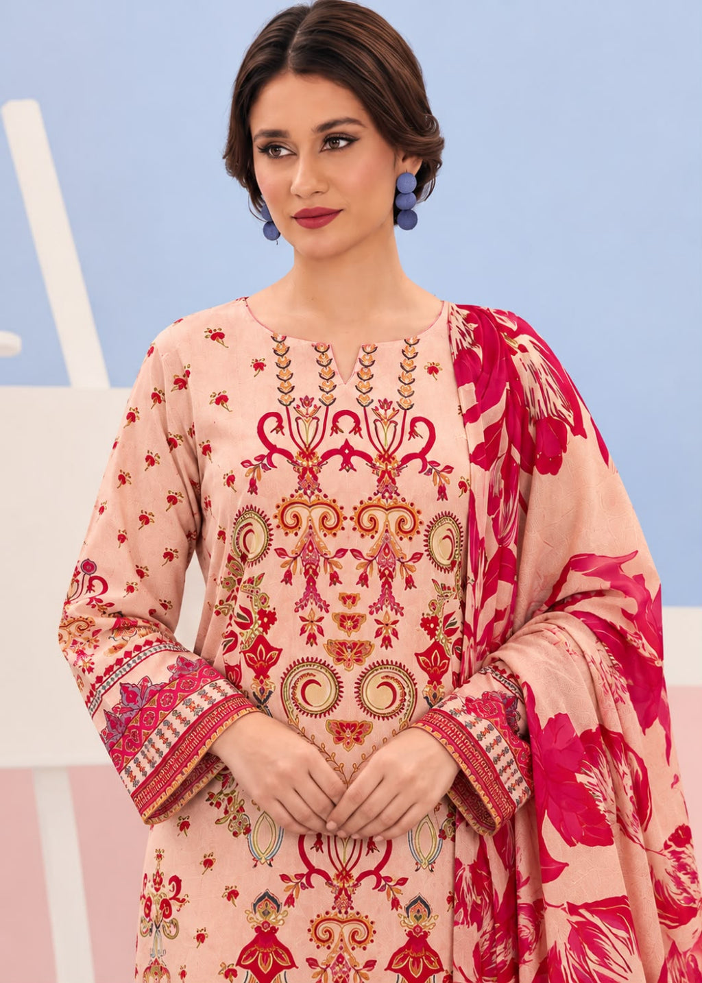 A woman modeling a peach-colored traditional embroidered kurta set with a matching printed dupatta. The kurta features intricate red, gold, and green embroidery on the chest and sleeves, with a vibrant pink and red floral pattern on the dupatta.
