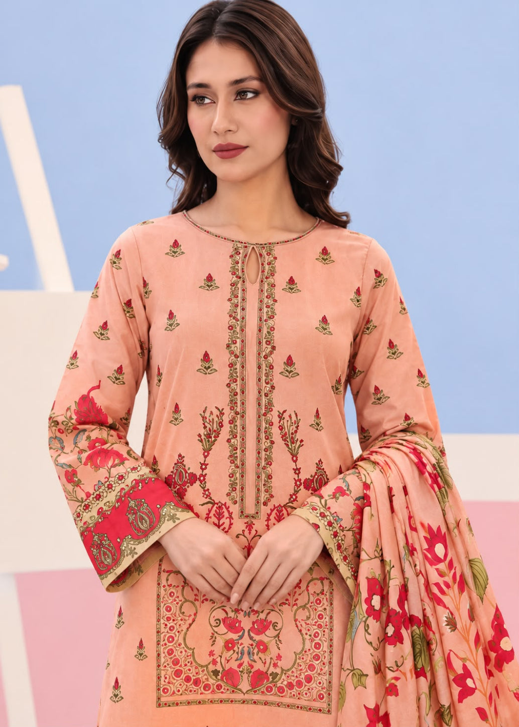A woman modeling a peach-colored traditional three-piece salwar kameez suit with red and gold floral prints and embroidery, paired with a matching dupatta.
