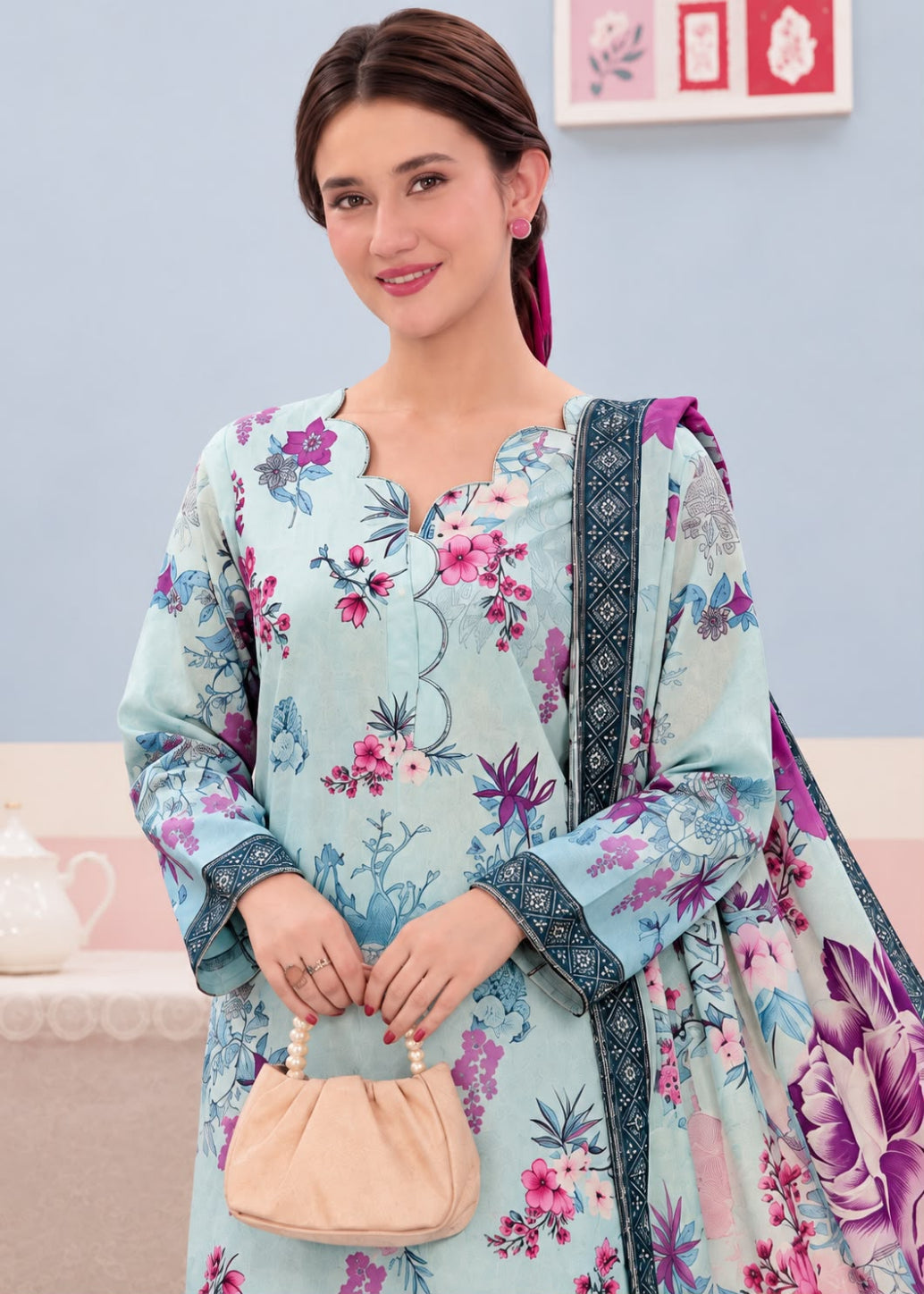 A woman modeling a light blue floral printed salwar kameez suit with a matching printed dupatta, holding a small beige handbag against a soft blue and pink background.
