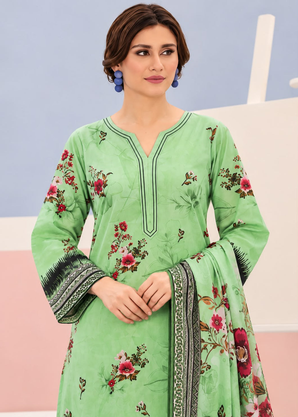 A woman wearing a light green traditional outfit with red floral prints and dark patterned borders on the sleeves and dupatta, set against a pastel studio background.
