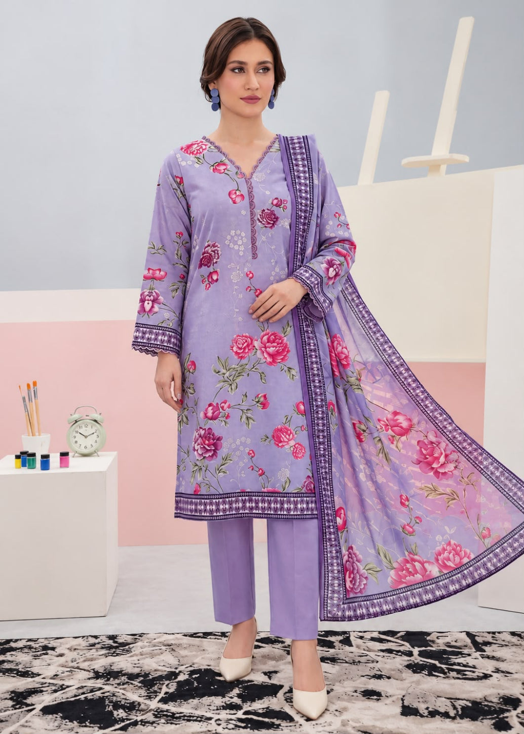 A woman models a three-piece lavender ethnic suit set featuring a floral printed kurta, matching trousers, and a sheer printed dupatta, set against a pastel studio background.