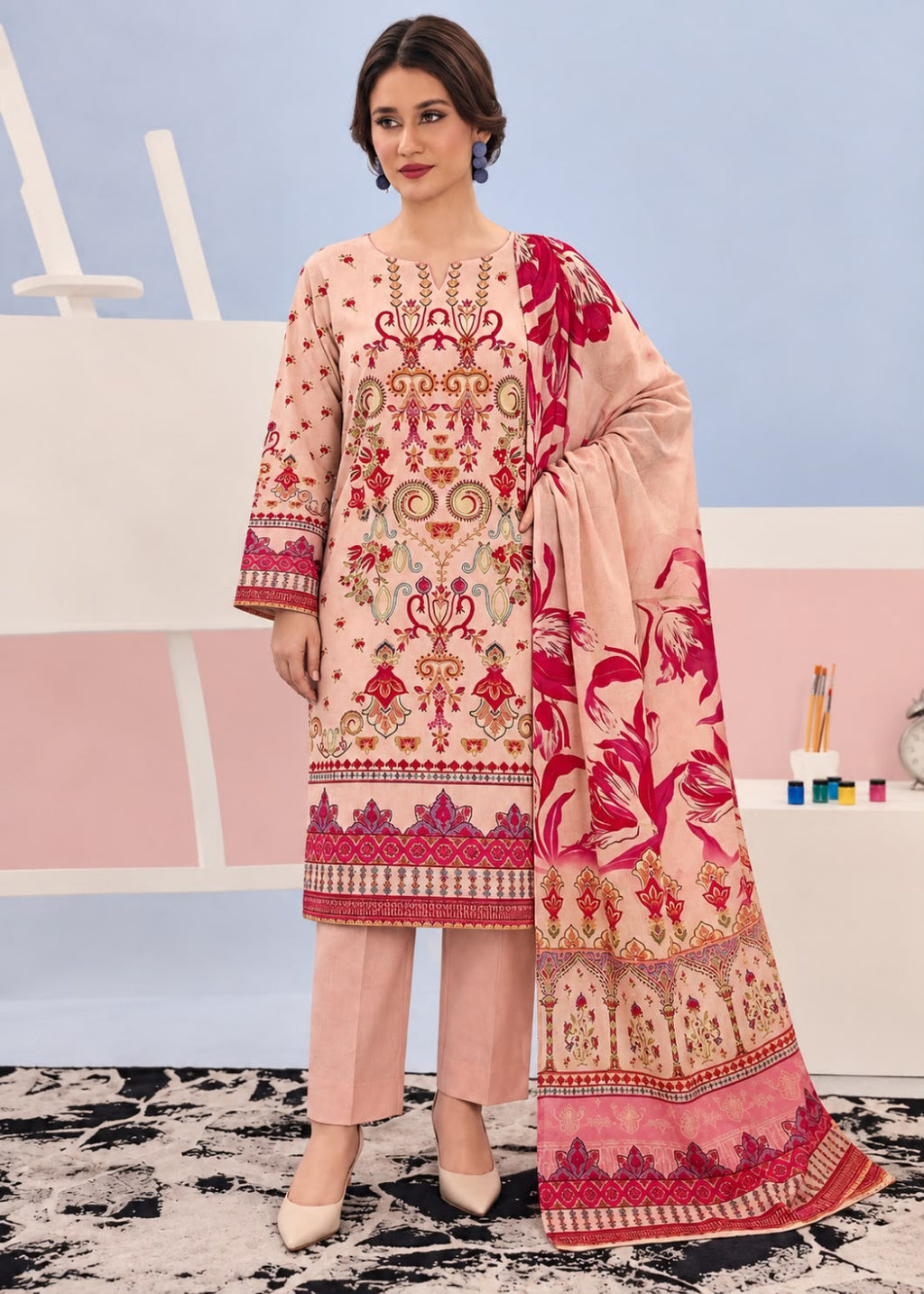 A woman modeling a traditional three-piece salwar kameez suit in light pink with intricate red and gold embroidery, paired with matching pink trousers and a large floral printed dupatta.