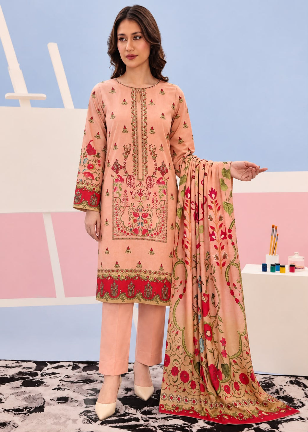A woman models a peach-colored three-piece ethnic suit with intricate red and gold floral embroidery, paired with matching trousers and a coordinating dupatta, set against a pastel studio backdrop.