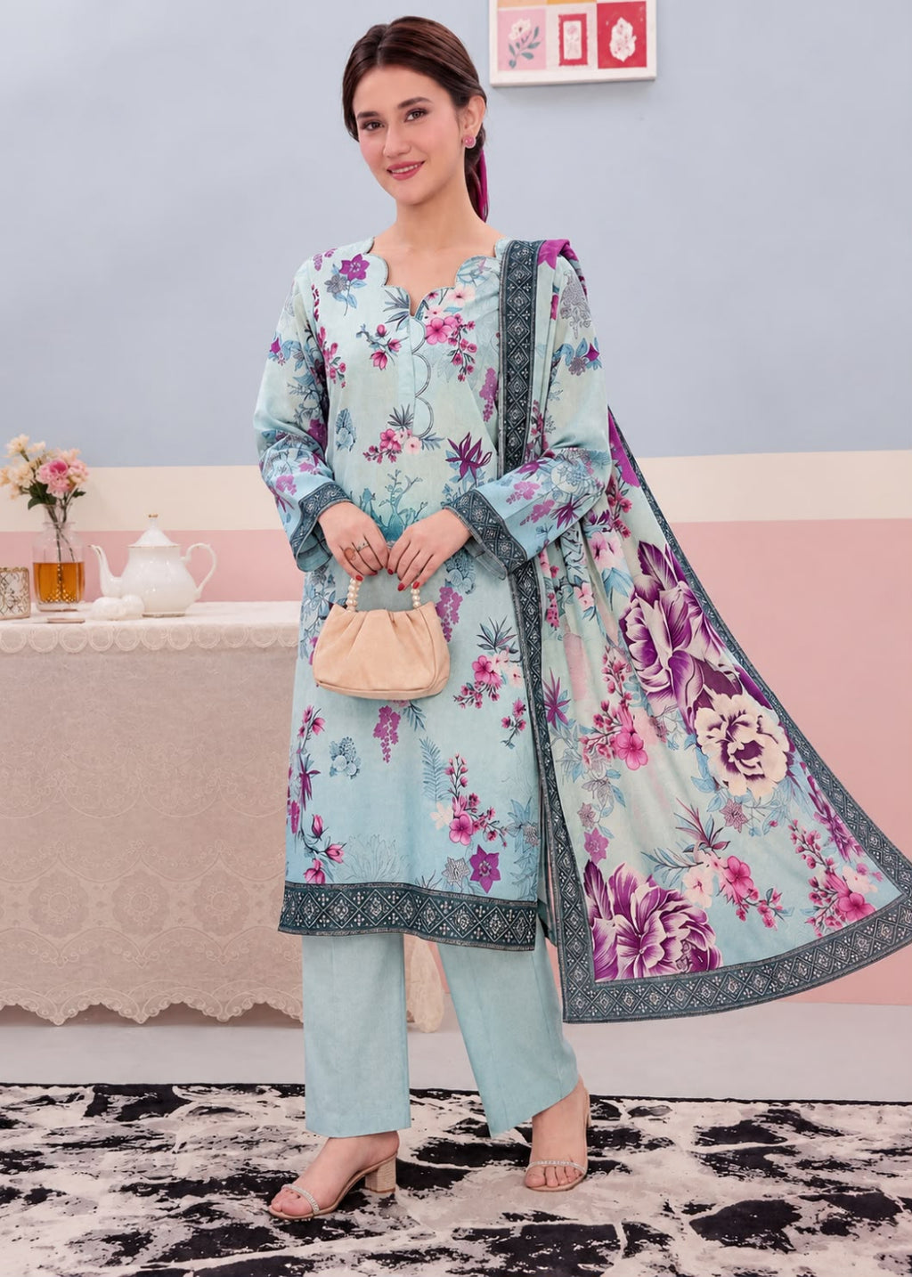 A woman models a light blue three-piece salwar kameez suit with a floral print, matching pants, and a dupatta, standing in a styled indoor setting.