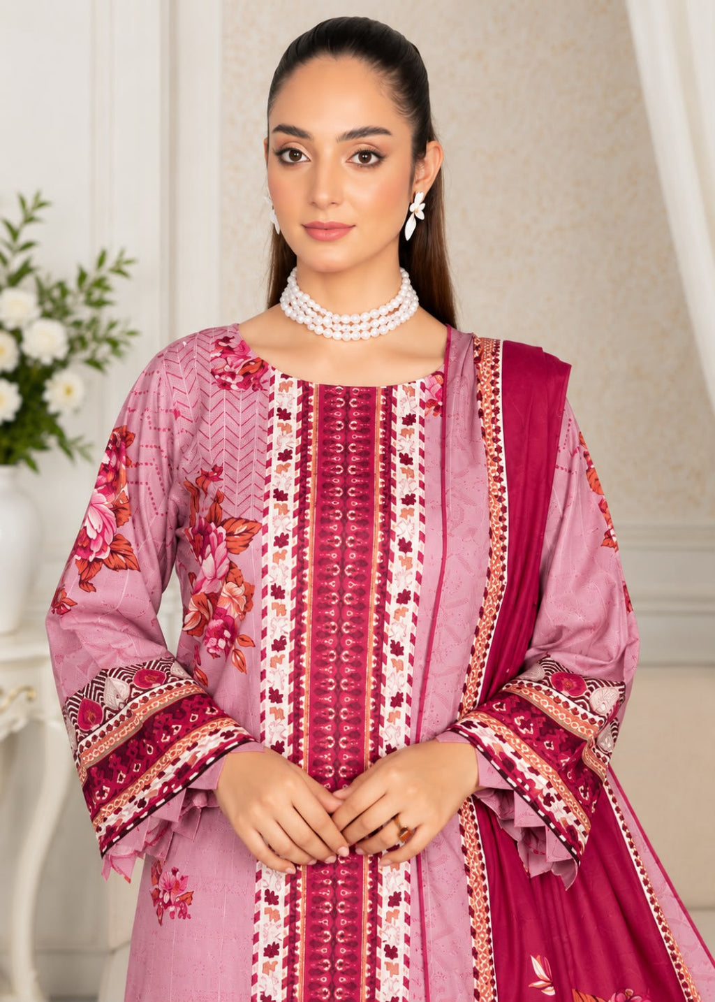 A woman modeling a traditional pink salwar kameez suit with floral embroidery, geometric patterns, and a matching magenta dupatta, set against an elegant indoor background.