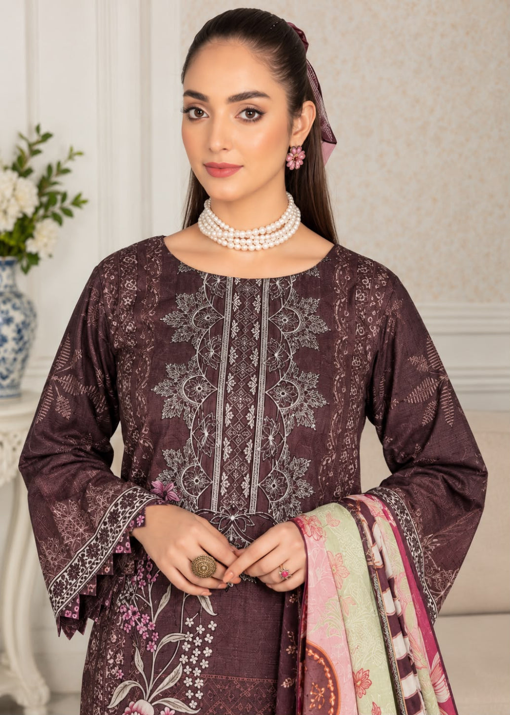 A woman modeling a dark plum embroidered kurta with matching printed dupatta, set against an elegant indoor background with a floral vase and cream-colored wall.
