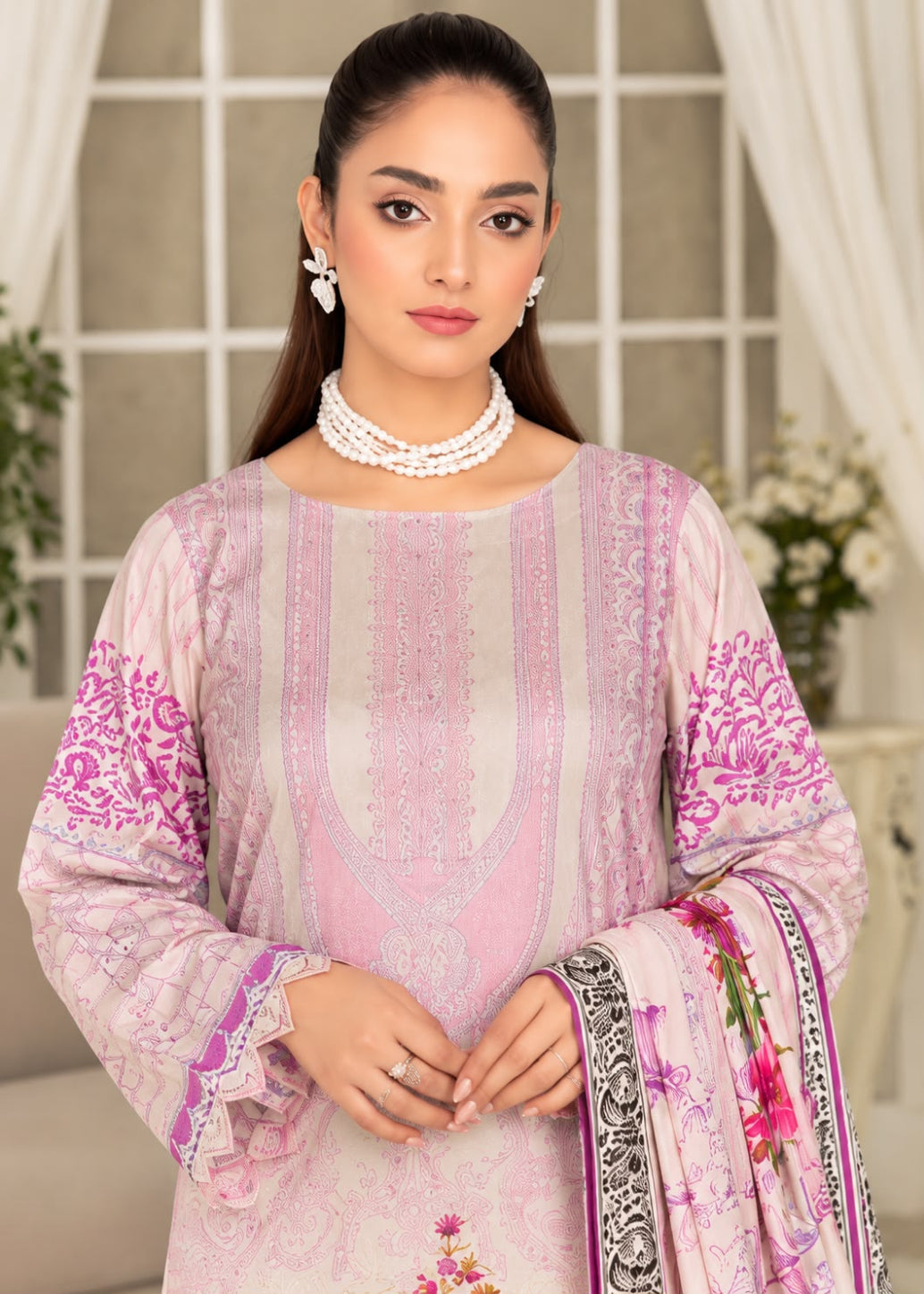 A woman modeling a light pink traditional South Asian salwar kameez suit with intricate silver embroidery, a matching pink floral dupatta, and white pearl jewelry, set against a soft-focus indoor background.
