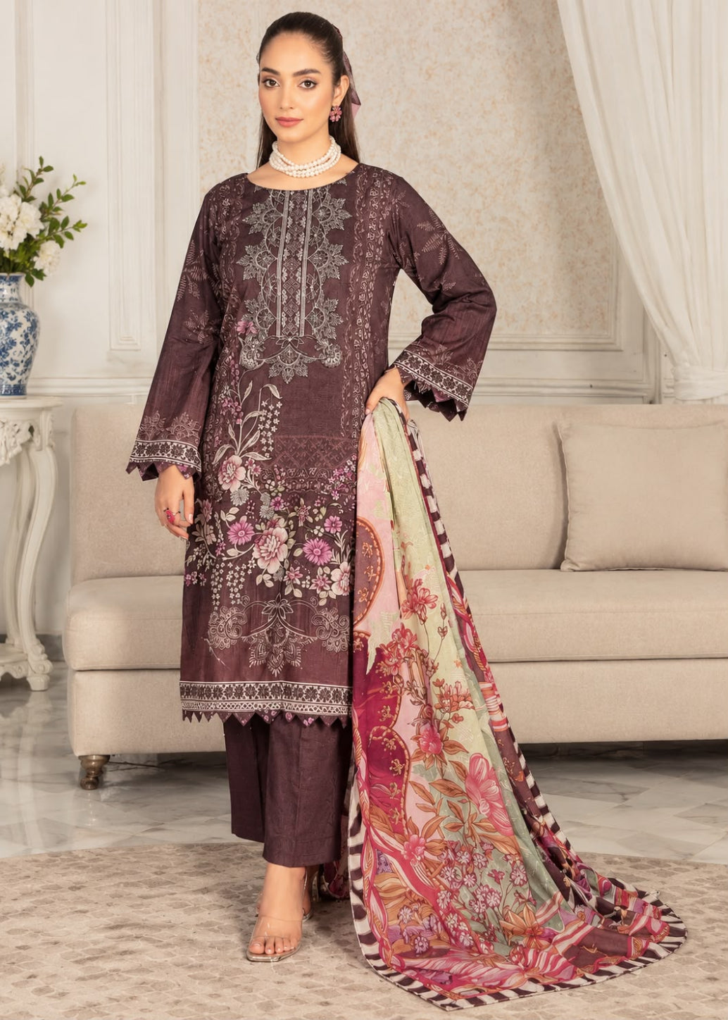 A woman models a three-piece traditional South Asian outfit consisting of a dark plum kurta with floral embroidery, matching trousers, and a large multicolored printed dupatta in a living room setting.