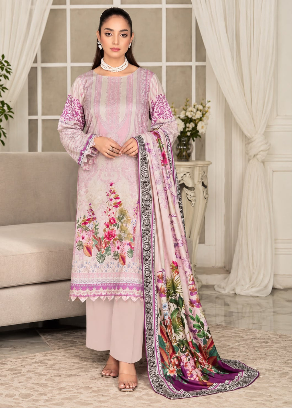 A woman models a traditional three-piece salwar kameez suit in light pink with floral embroidery and prints, paired with a matching printed dupatta, in an elegant indoor setting.