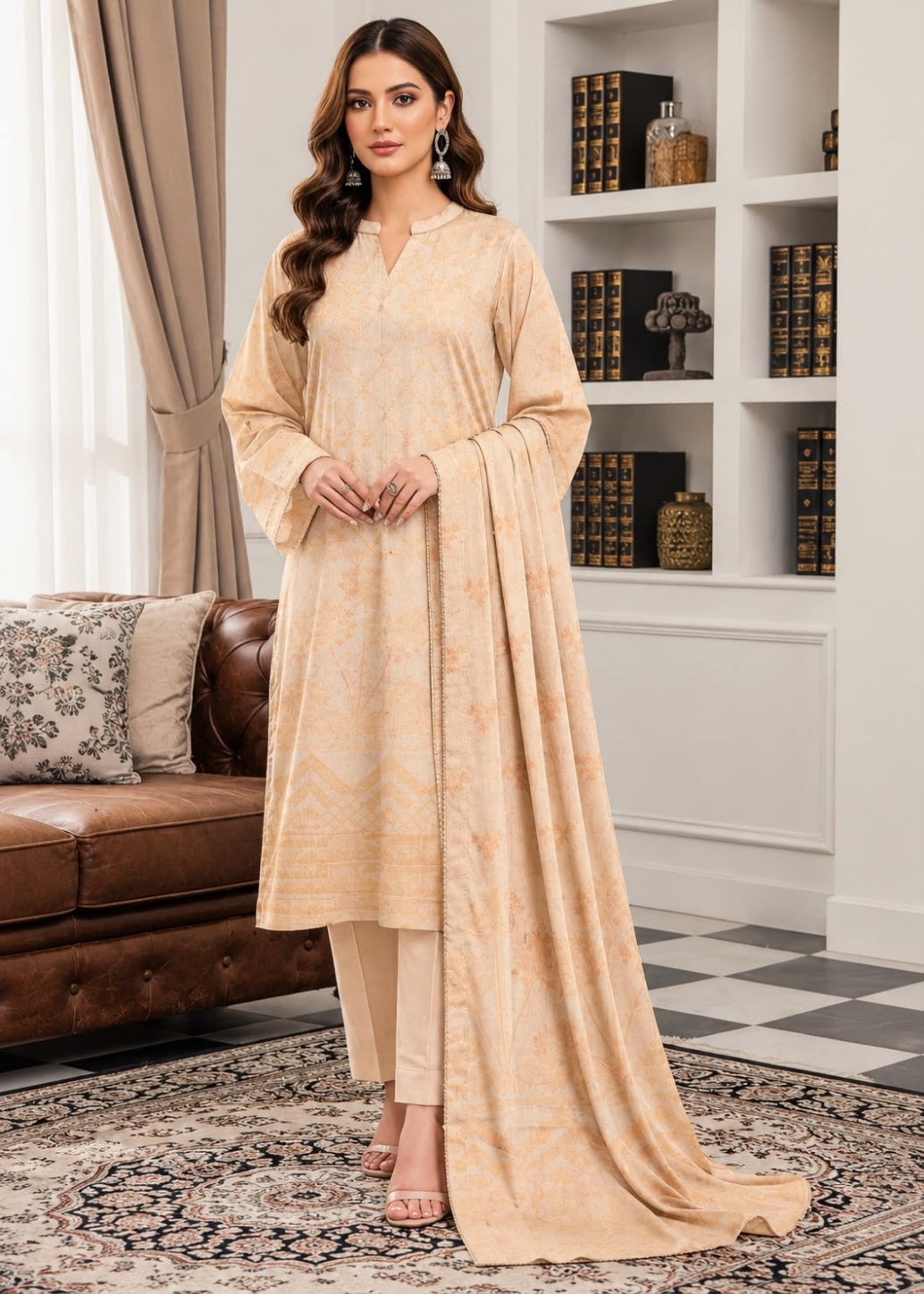 A woman models a traditional three-piece salwar kameez suit in a warm beige/cream color with a subtle marbled pattern, featuring a long kurta, matching trousers, and a draped dupatta, set against an elegant indoor background with a bookshelf and leather sofa.