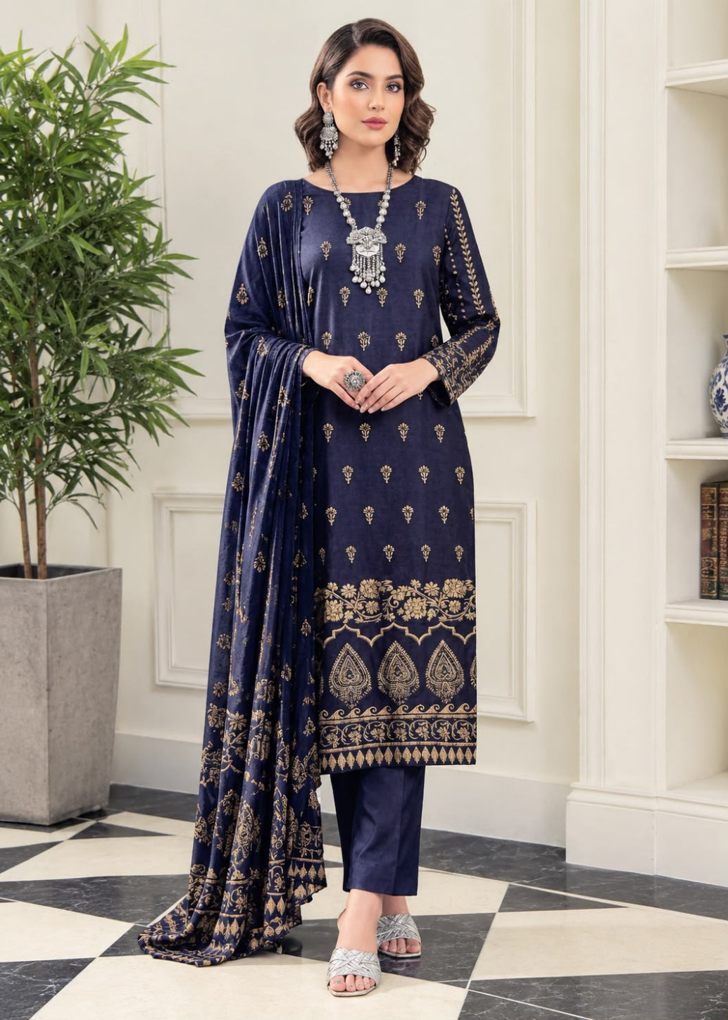 A woman models a navy blue ethnic three-piece suit with gold embroidery, featuring a kurta, trousers, and a dupatta, set against an elegant indoor background with a potted plant and white paneled walls.