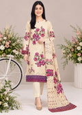 A woman models a traditional three-piece salwar kameez suit set in cream beige, featuring vibrant magenta and green floral prints, a patterned purple border, and a matching dupatta with floral motifs and a patterned border. The background is a soft beige studio setting decorated with floral arrangements and a white bicycle prop.
