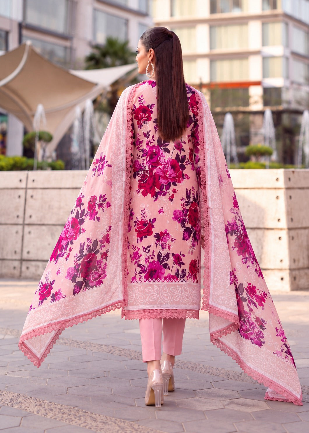 A woman viewed from behind wearing a pink traditional outfit with a large floral print dupatta and matching pants, walking outdoors in an urban plaza setting.