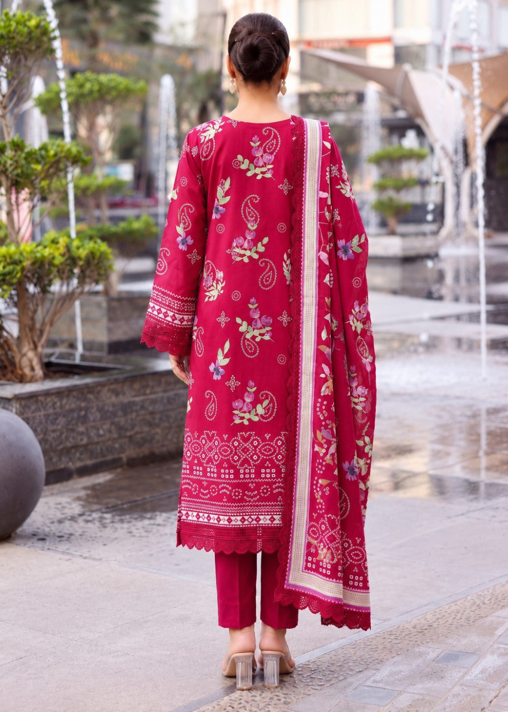 A woman viewed from behind wearing a bright magenta-red three-piece salwar kameez suit with intricate multicolor floral and paisley embroidery, standing in an outdoor courtyard with water fountains in the background.