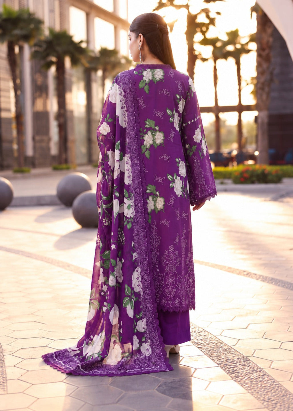 A woman is shown from behind wearing a purple traditional outfit with a matching floral dupatta in an outdoor plaza at sunset.