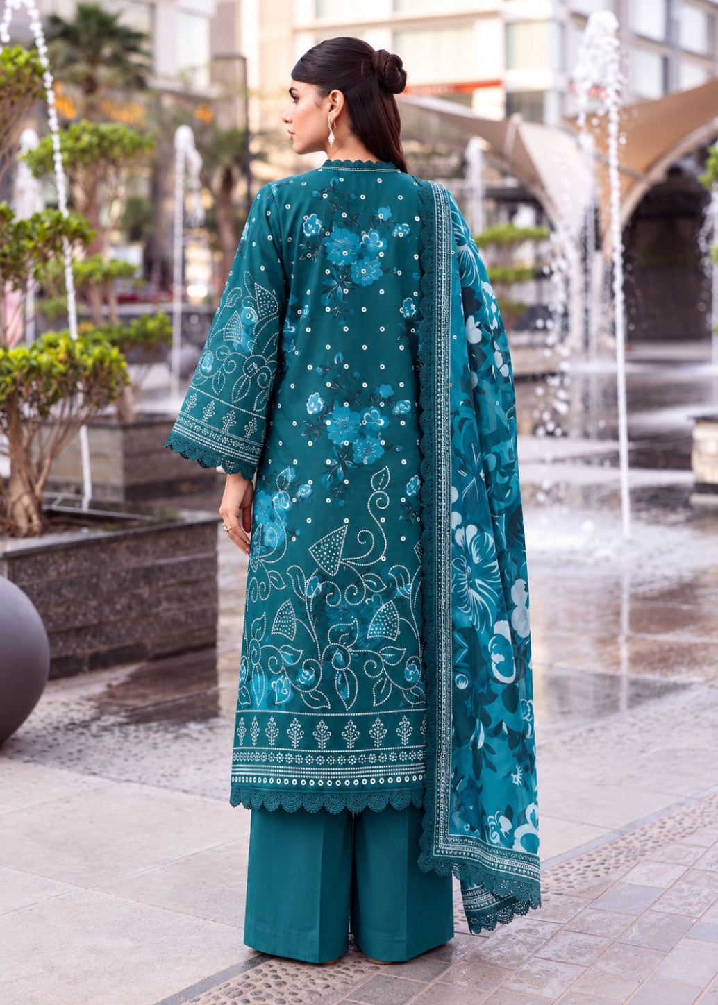 A woman stands outdoors in front of water fountains, wearing a teal embroidered kurta with matching wide-leg trousers and a coordinating dupatta. The outfit features light blue and white floral patterns and ornate border embroidery.