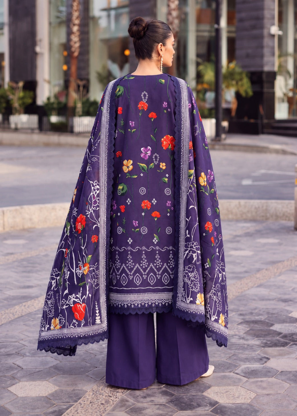 A woman stands outdoors on a paved walkway wearing a dark purple traditional outfit with colorful floral embroidery and a matching embroidered dupatta.