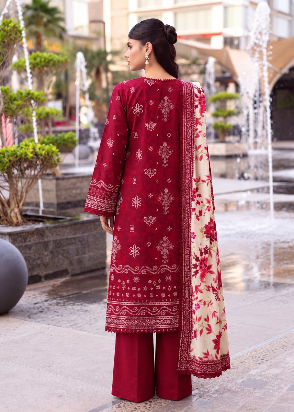 A woman stands outdoors in front of a fountain plaza, wearing a deep red three-piece traditional outfit with intricate gold embroidery, matching wide-leg pants, and a cream dupatta with red floral patterns.