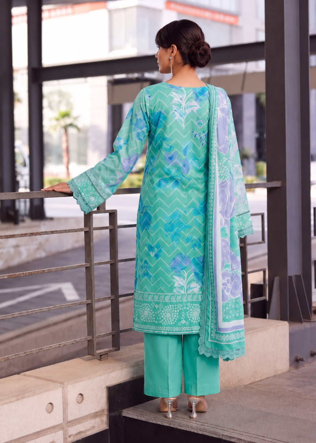 A woman stands on outdoor concrete steps beside a metal railing, wearing a mint green traditional outfit with a matching printed dupatta. The outfit has blue and white geometric and floral patterns, paired with solid mint green pants.