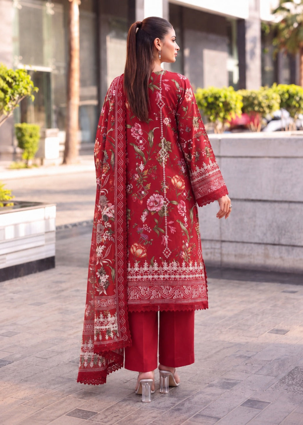 A woman wearing a red three-piece traditional outfit with floral embroidery and a matching dupatta, photographed from behind in an outdoor urban setting.