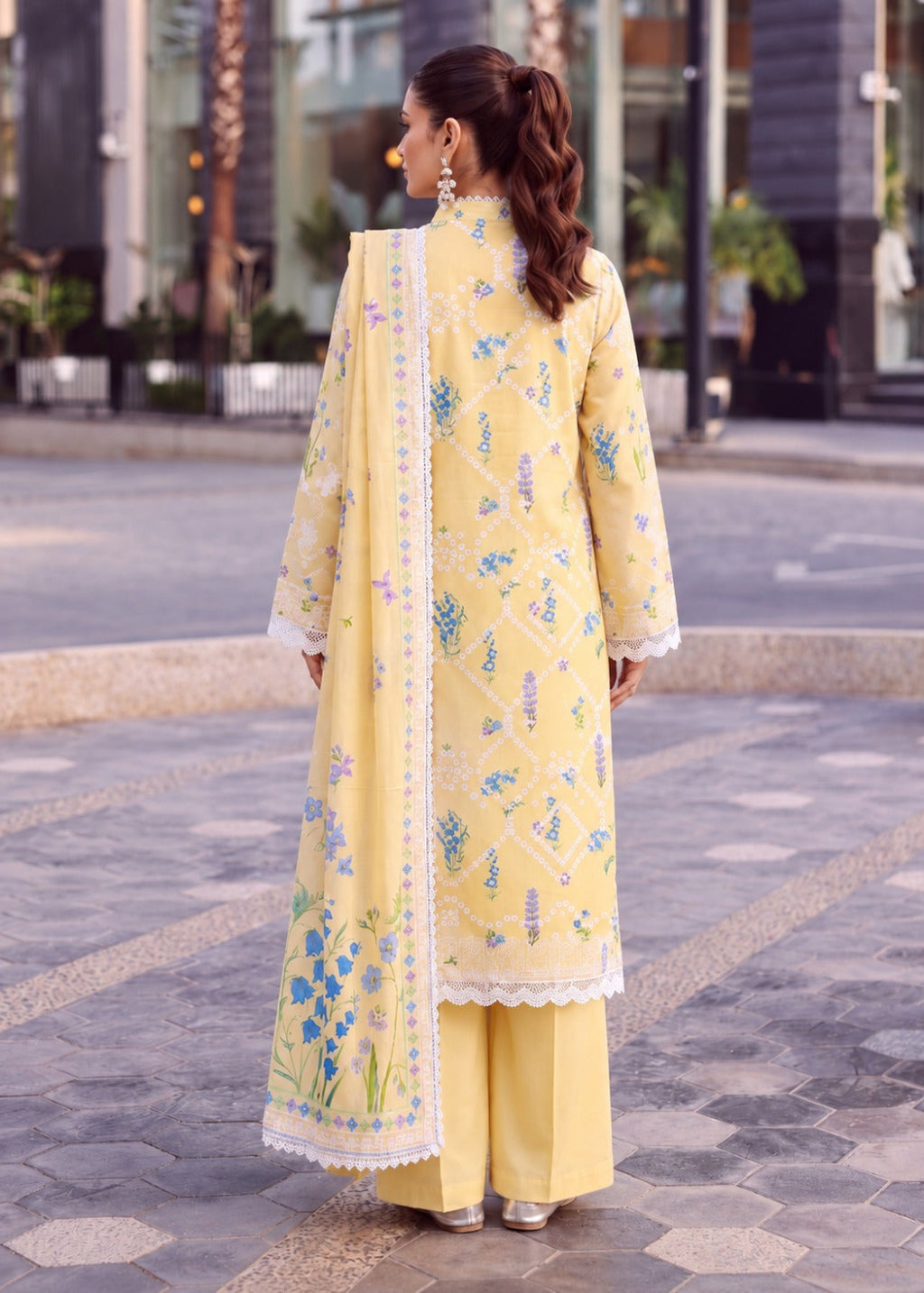 A woman stands outdoors on a paved street wearing a light yellow traditional outfit with floral embroidery, a matching long scarf, and wide-leg pants. The photo is taken from behind, showing the back of the kurta, scarf, and trousers.
