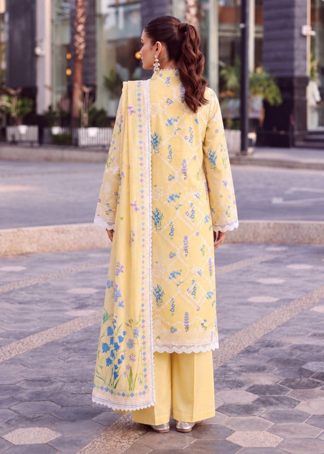 A woman stands outdoors on a paved street wearing a light yellow traditional outfit with floral embroidery, a matching long scarf, and wide-leg pants. The photo is taken from behind, showing the back of the kurta, scarf, and trousers.