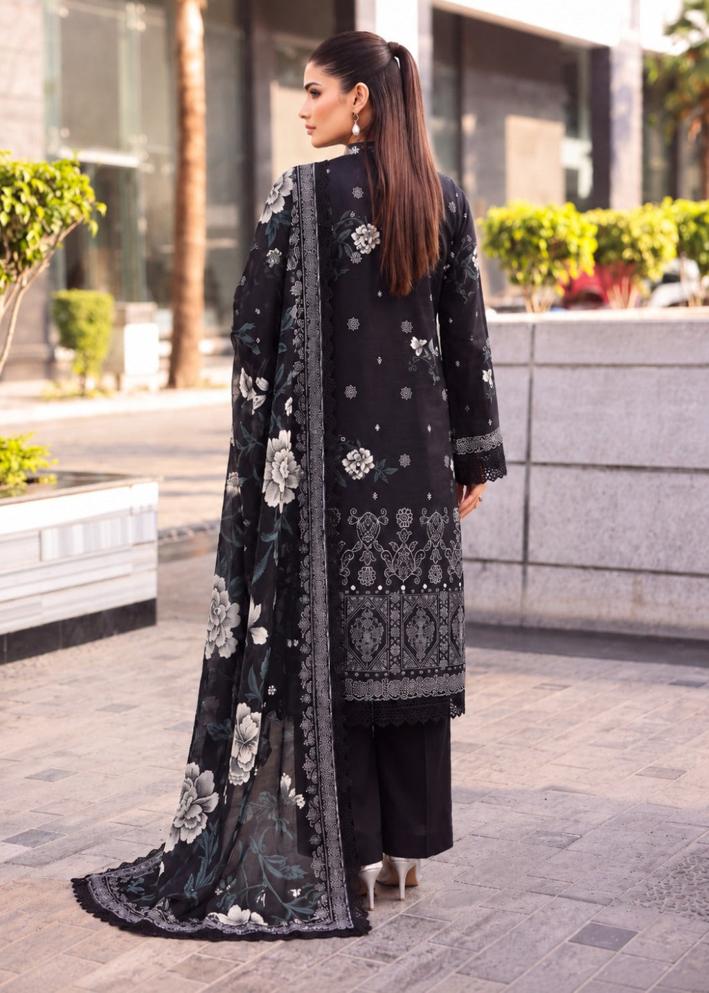A woman wearing a black traditional outfit with intricate silver floral embroidery and a matching dupatta, photographed outdoors on a paved walkway.