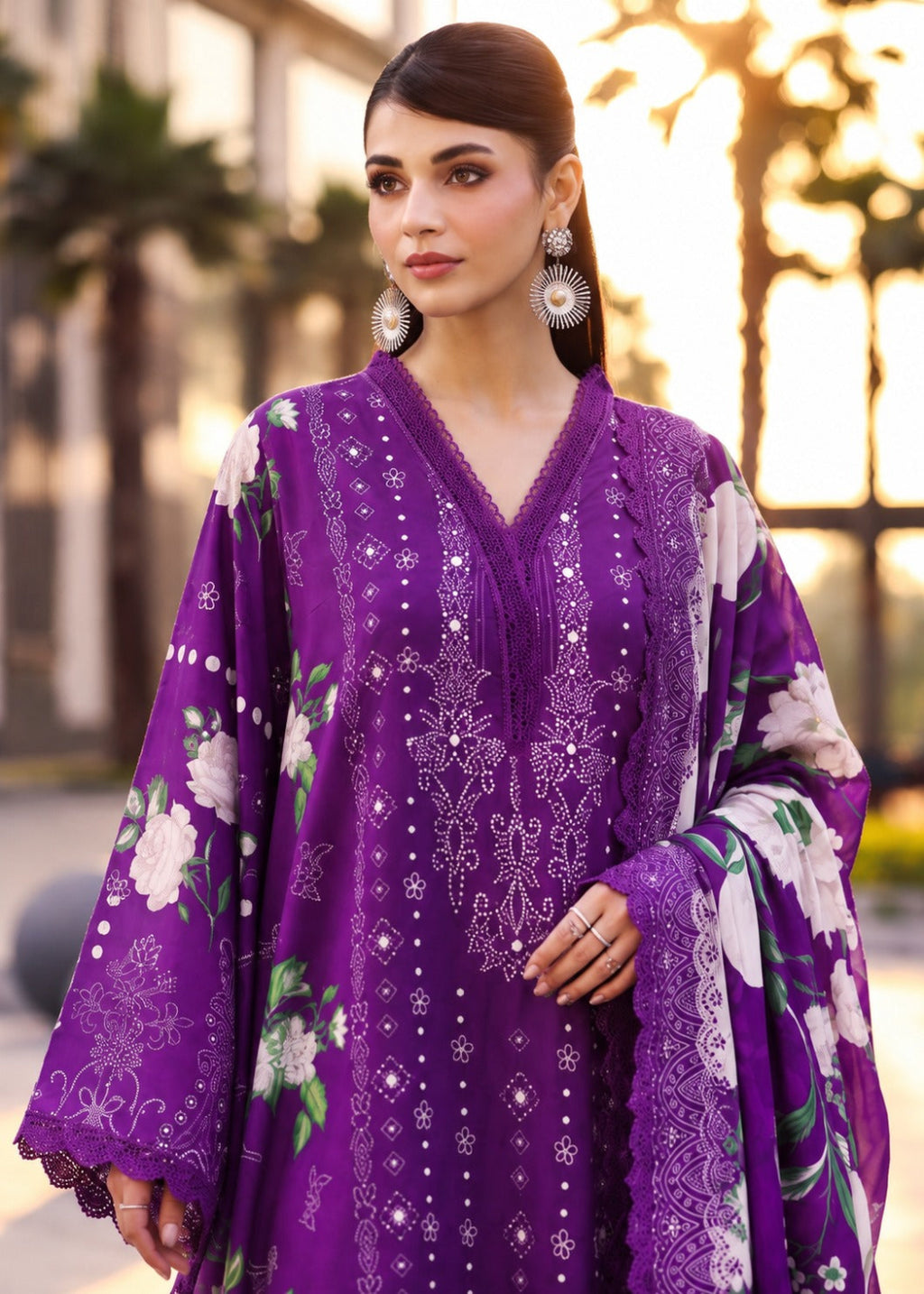 A woman wearing a purple embroidered traditional outfit with floral prints and silver-toned jewelry, photographed outdoors with blurred palm trees and warm sunlight in the background.