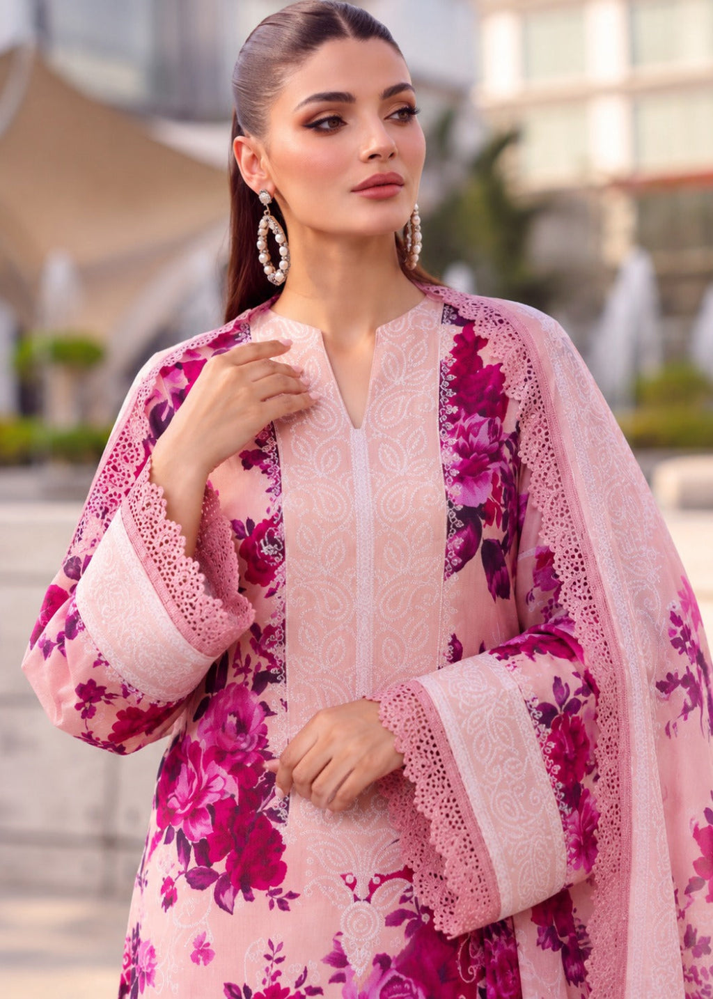 A woman wearing a pink floral traditional outfit with matching sheer dupatta, lace-trimmed sleeves, and a lace front, photographed outdoors with a softly blurred urban background.