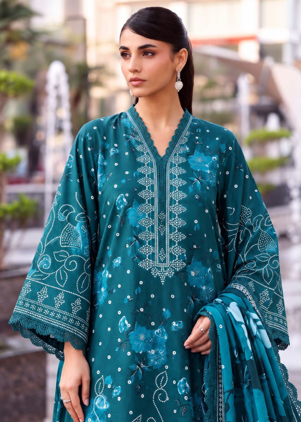 A woman wearing a teal green embroidered traditional kurta set with a matching dupatta, standing in an outdoor garden setting.