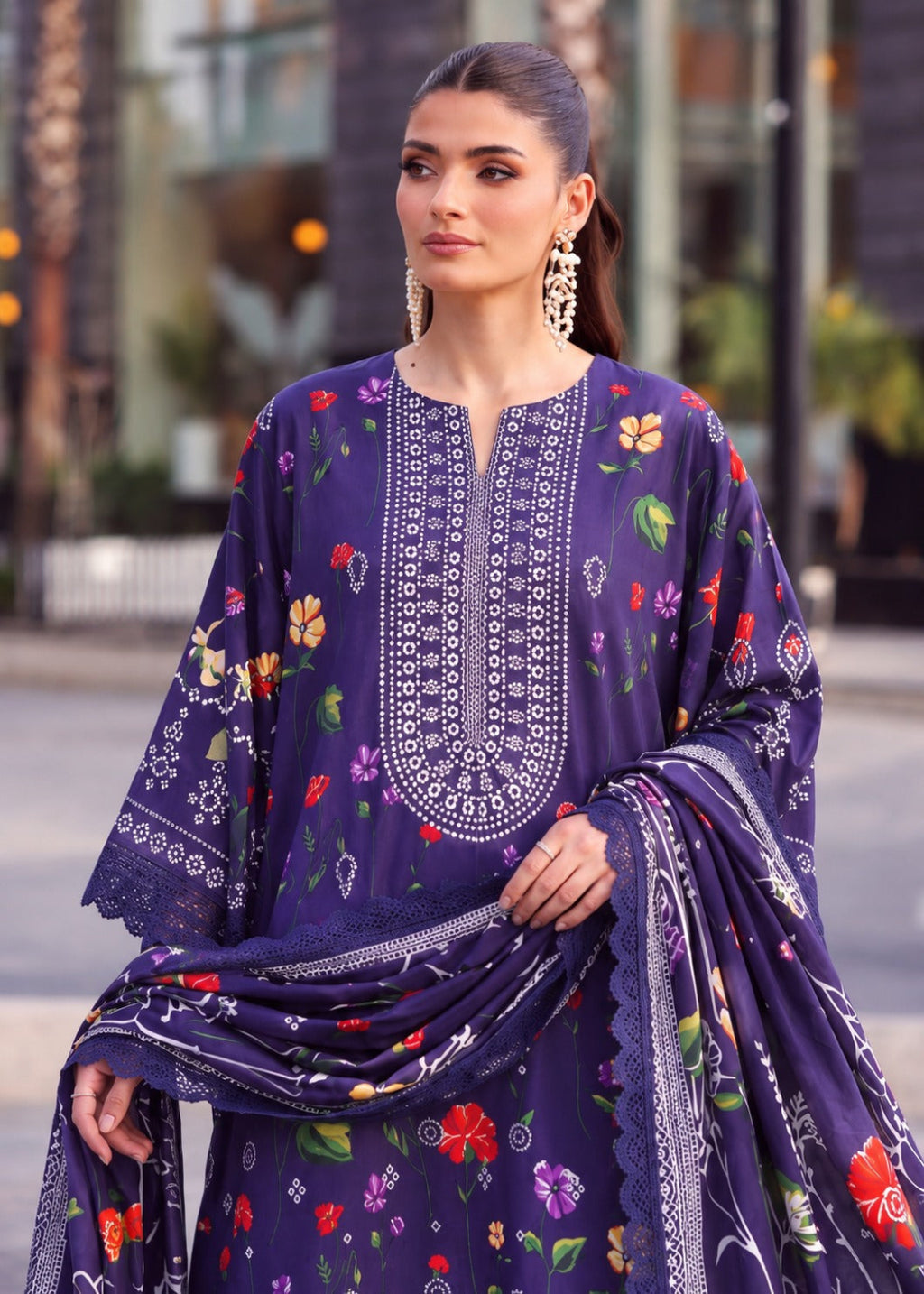 A woman wearing a purple floral printed traditional outfit with white embroidery and a matching dupatta, photographed outdoors against a softly blurred urban background.