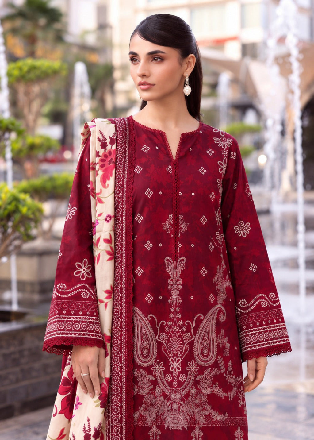 A woman wearing a deep maroon traditional kurta set with intricate cream paisley and floral embroidery, paired with a matching cream dupatta featuring red floral prints, set against a blurred outdoor background with fountains.