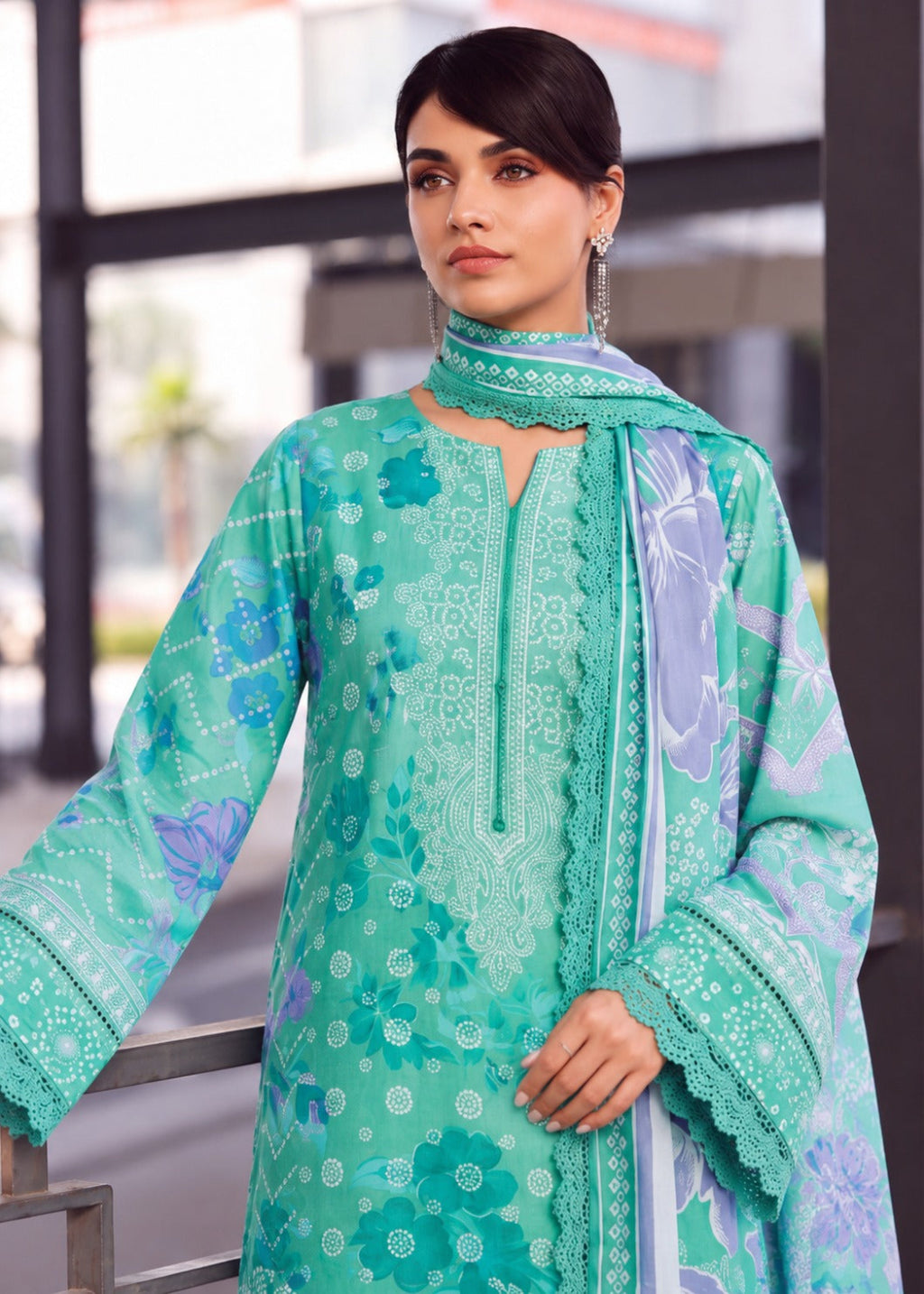 A woman modeling a mint green ethnic kurta set with floral prints, featuring a matching dupatta and embroidered details, set against a blurred modern building background.