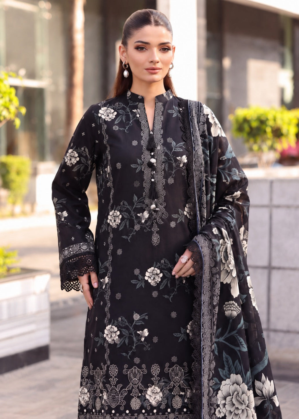 A woman modeling a black three-piece ethnic suit with intricate silver and white floral embroidery, featuring a long kurta, matching trousers, and a sheer dupatta.
