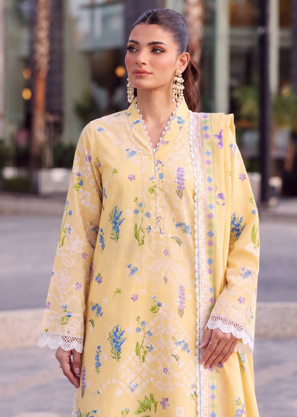 A woman wearing a light yellow embroidered traditional outfit with floral patterns and lace trim, standing outdoors in a softly blurred urban setting.