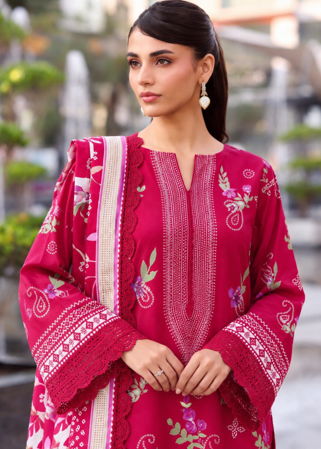 A woman wearing a bright pink embroidered traditional kurta with a matching dupatta, featuring floral and paisley patterns, set against a softly blurred outdoor background.